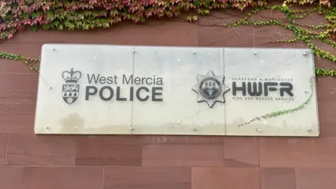 BBC A sign on a red brick wall that reads "West Mercia Police" on the left hand side and "Hereford and Worcester Fire and Rescue" on the right. There are green and red leaves at the top of the image. 