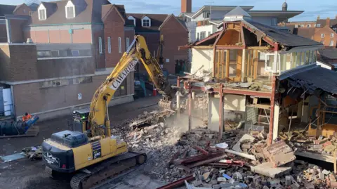 BBC A large yellow digger with a claw is pointing towards a half-demolished building. There are bricks, rubble and wood all over the floor.