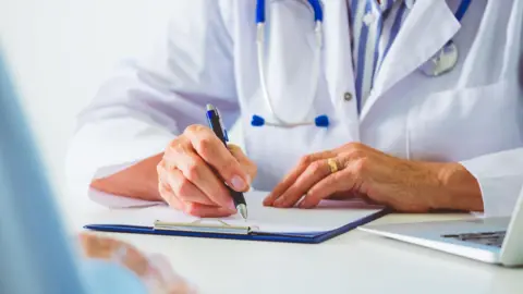 Getty Images A stock image of a medical professional in a white doctor's coat writing on a clip board