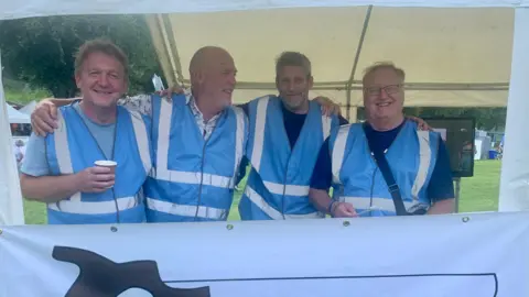 Shropshire Council Four men are standing in a large tent, all wearing blue vests with white reflective strips. They are all smiling and laughing, with one holding a coffee cup.