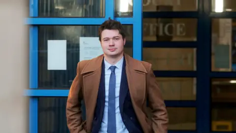 SWNS A man in a light brown overcoat with a navy suit jacket, blue tie and light blue shirt underneath. He has short brown hair. He is facing the camera with his hands in his pocket. Behind him is a blue door that is partially open