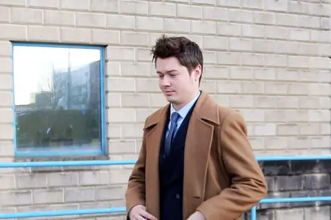 SWNS A man in a light brown overcoat with a navy suit jacket, blue tie and light blue shirt underneath. He has short brown hair and has a slight grin. Behind him is a wall of grey bricks with a window.