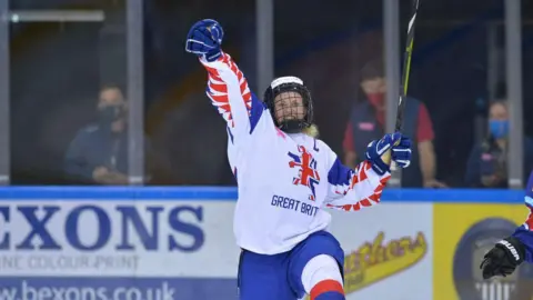 Ice Hockey UK A woman in a white ice hockey jersey with Great Britain and a stylised lion printed on the front, wearing a blue helmet and holding an ice hockey stick in her left hand. She has one hand in the air and there are advertising hoardings behind her